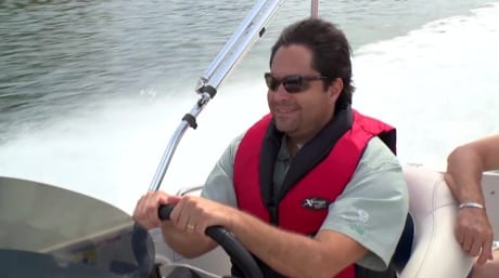A man drives a boat while wearing a life jacket.
